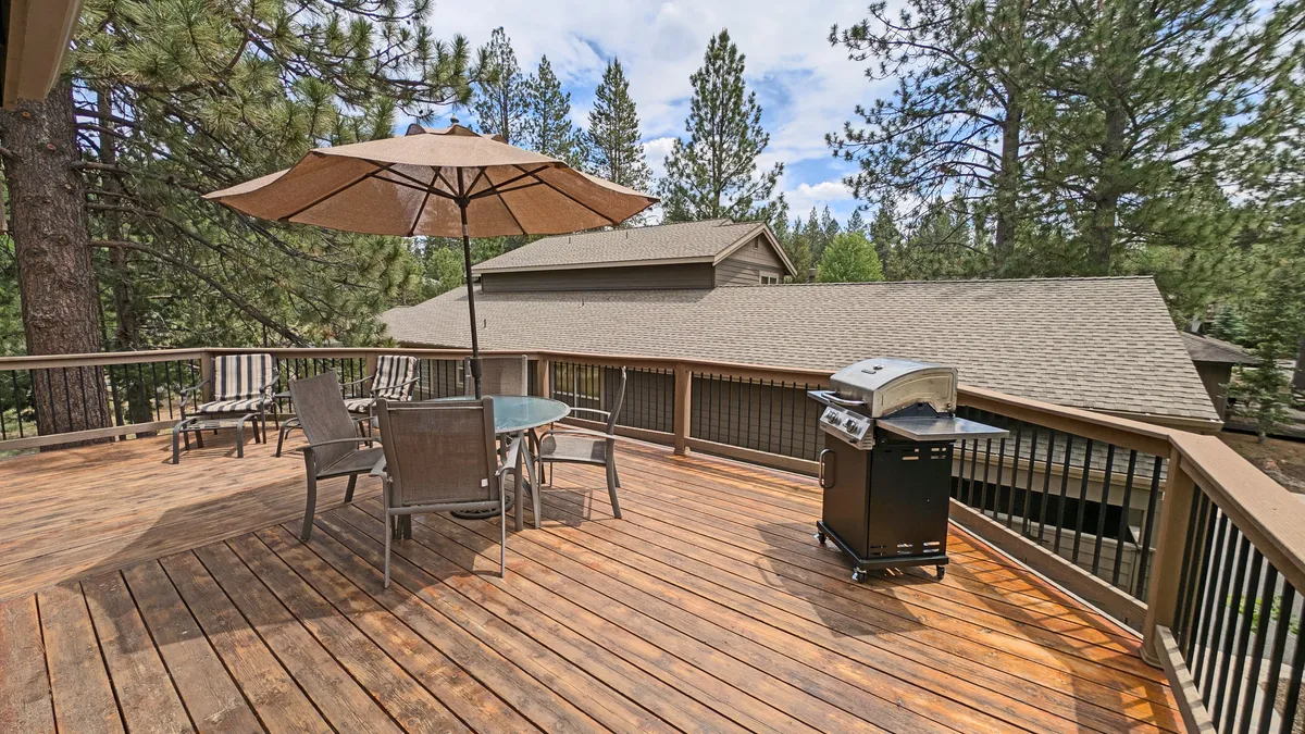 An inviting outdoor dining area sits on a wooden deck, featuring a large grill, table with chairs, and a sun umbrella. The space is designed for barbecuing, al fresco meals, and group gatherings, with the lush forest providing a scenic backdrop.