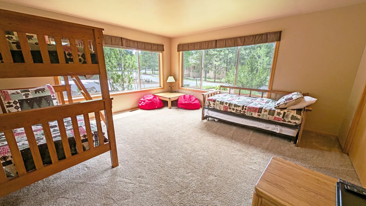 A vibrant, playful kids' bunk room with bunk beds, bean bags, a bed with patterned bedding, large windows, and a side table with a lamp. The room exudes a lively, cozy atmosphere perfect for play, sleep, and bonding.