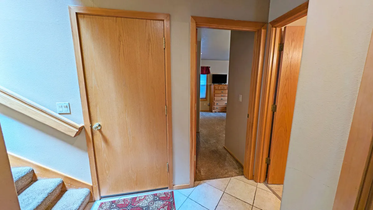 A bright, welcoming entryway with wooden doors, a staircase, tile flooring, and an area rug. The image conveys both arrival and departure—guests moving between private and communal spaces, possibly on their way out for a bike ride or returning from a day on the river or in the snow.