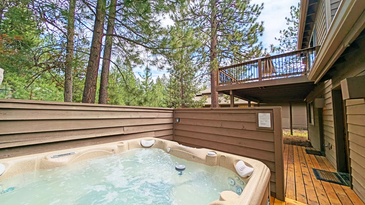 This image captures a spacious wooden deck nestled among tall pine trees, with a bubbling hot tub as the focal point. The setting is tranquil, inviting guests to relax in the warm water while surrounded by nature. Comfortable seating is visible, and the deck seamlessly blends into the forest, suggesting privacy and serenity.
