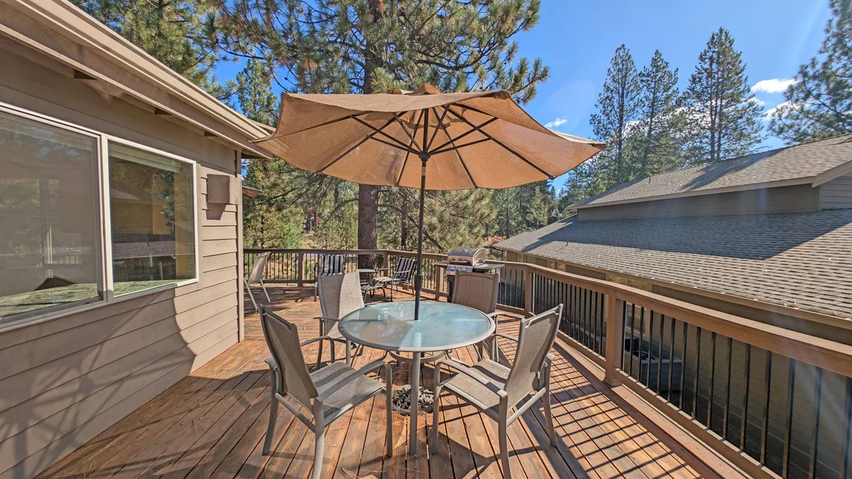 An exterior patio scene with a spacious outdoor dining table beneath a large umbrella, nestled among lush trees. The setup suggests opportunities for al fresco meals, casual gatherings, and enjoying the peaceful outdoor environment with family or friends.