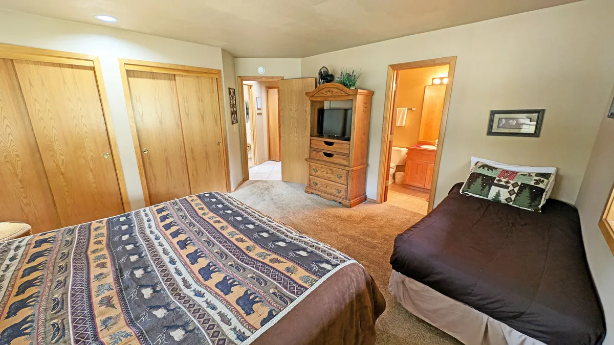 A cozy bedroom featuring a large bed with a patterned blanket, a wooden dresser, a small bed on the side ideal for a child, and closet doors. The atmosphere is welcoming and versatile, suitable for families or couples with children.