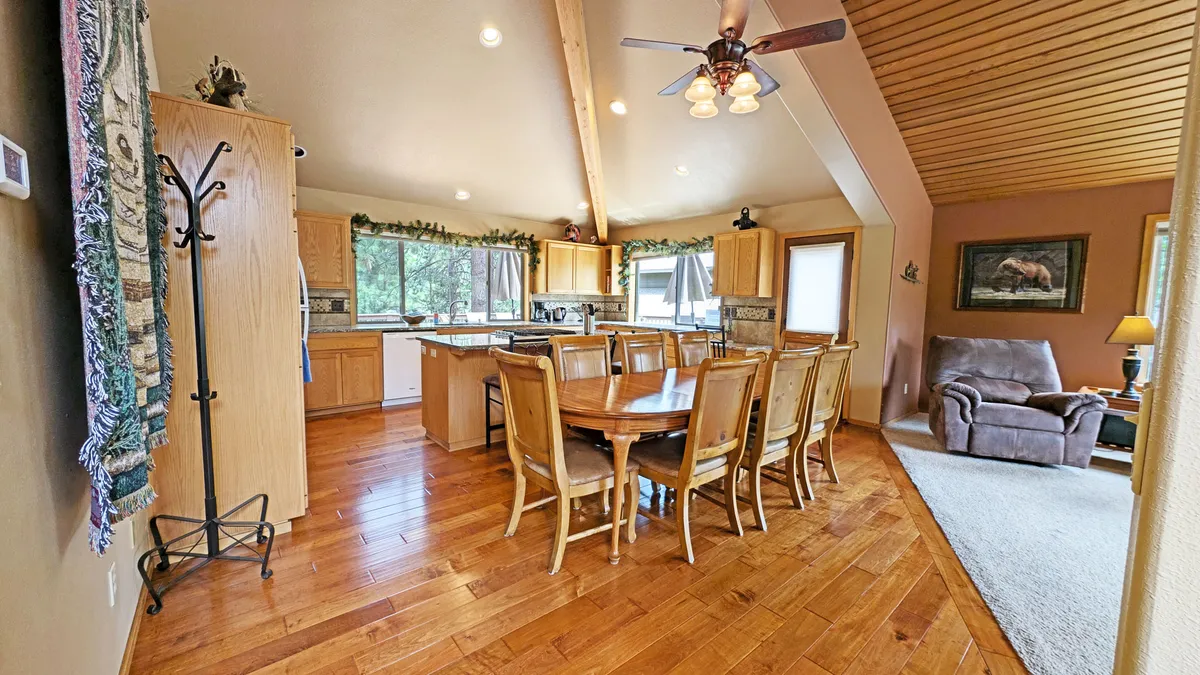 This image captures a wide view of the open-plan area, seamlessly linking the dining table, kitchen, and living room seating. Wooden beams overhead and large windows infuse the space with warmth and natural light, while the thoughtful layout encourages easy movement and togetherness.