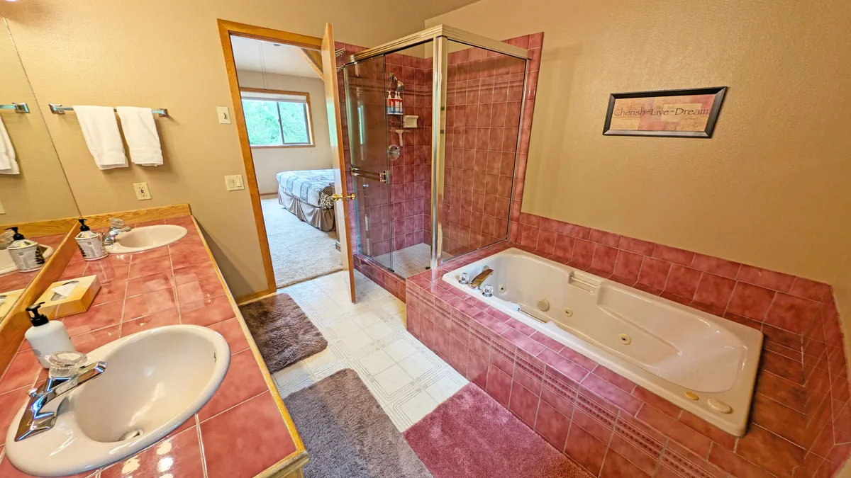 This image showcases a spacious bathroom complete with a double sink vanity, a glass shower enclosure, a whirlpool soaking bathtub, and a window that lets in plenty of natural light. The serene and comfortable mood invites relaxation and private moments of self-care.