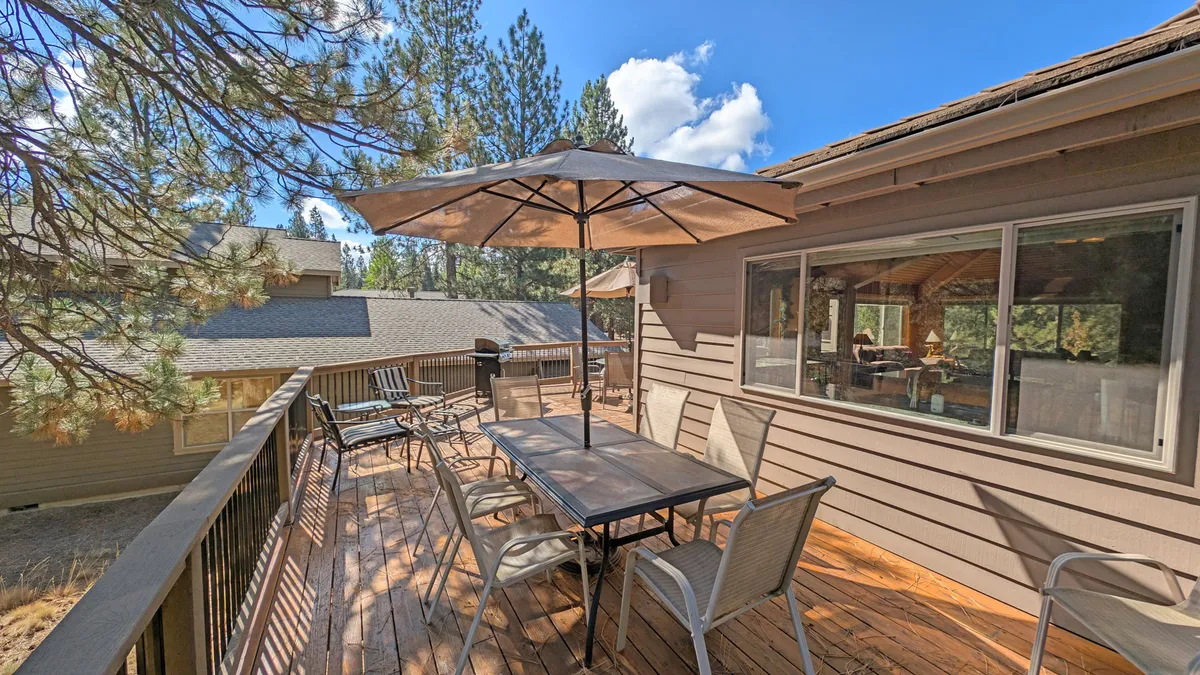 An inviting exterior patio with an outdoor dining table shaded by an umbrella, set against a background of lush trees. The space is ideal for al fresco meals, morning coffee, or evening conversations under the stars, seamlessly connecting the home to its natural surroundings.